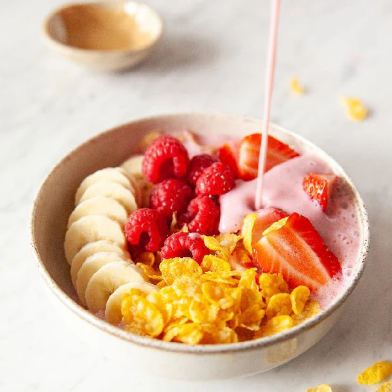Healthy breakfast bowl with organic gluten-free cornflakes, fresh strawberries, raspberries, sliced banana, and pouring pink yogurt smoothie